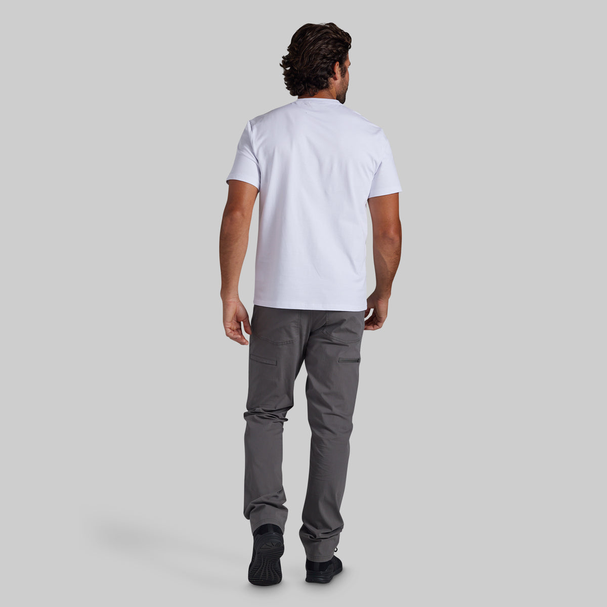 A man viewed from behind wearing a white short-sleeve t-shirt and dark gray pants, standing against a solid light gray background.