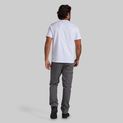 A man viewed from behind wearing a white short-sleeve t-shirt and dark gray pants, standing against a solid light gray background.