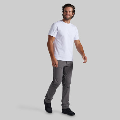 A man modeling a white short-sleeve crewneck t-shirt and gray pants against a solid gray background.