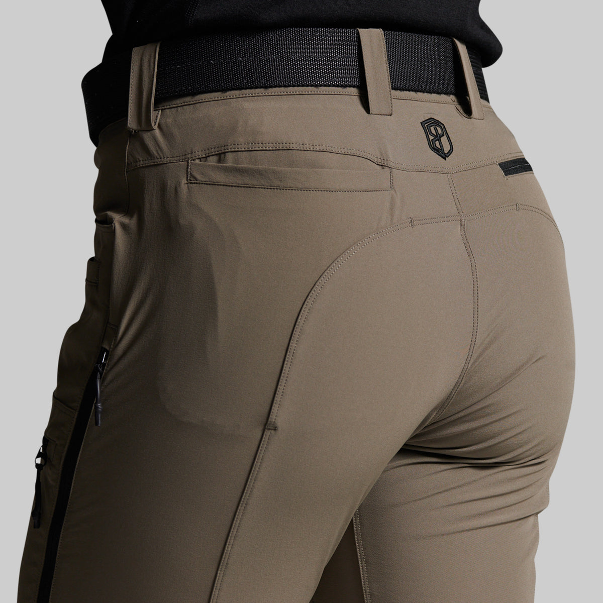 Close-up back view of a person wearing storm grey outdoor pants with a black belt, showing the rear pockets and a small shield logo near the waistband.