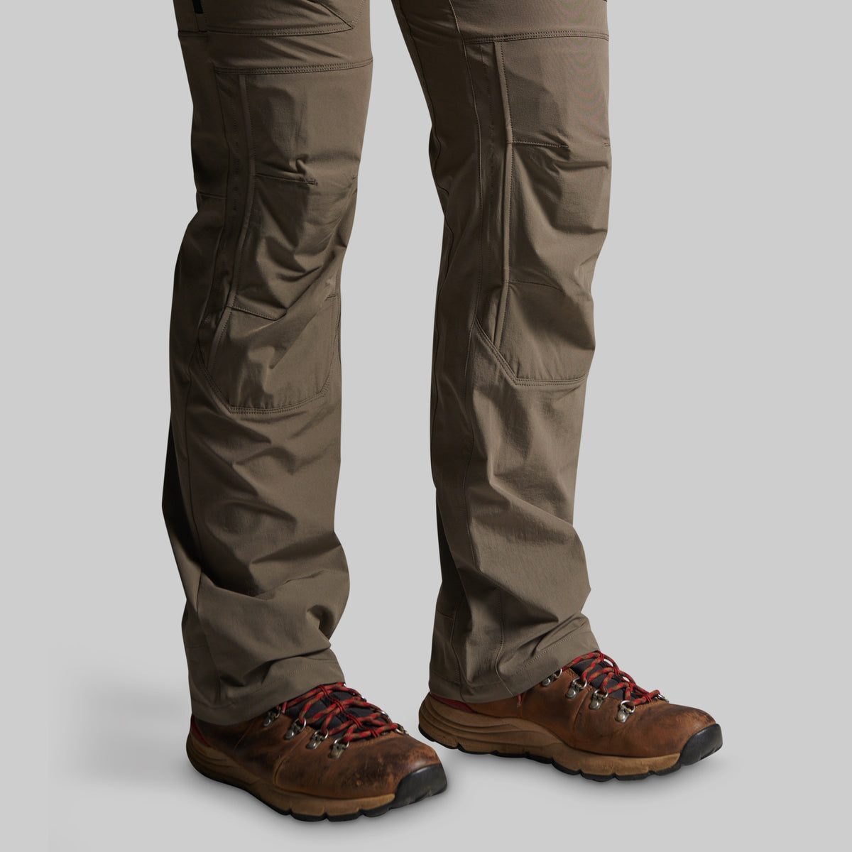 A close-up of a person's legs wearing brown hiking boots and storm grey cargo pants against a solid gray background.