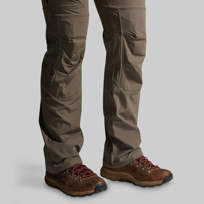 A close-up of a person's legs wearing brown hiking boots and storm grey cargo pants against a solid gray background.