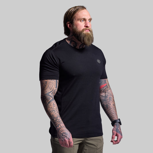 Man wearing a black t-shirt with visible tattoos on arms against a gray background