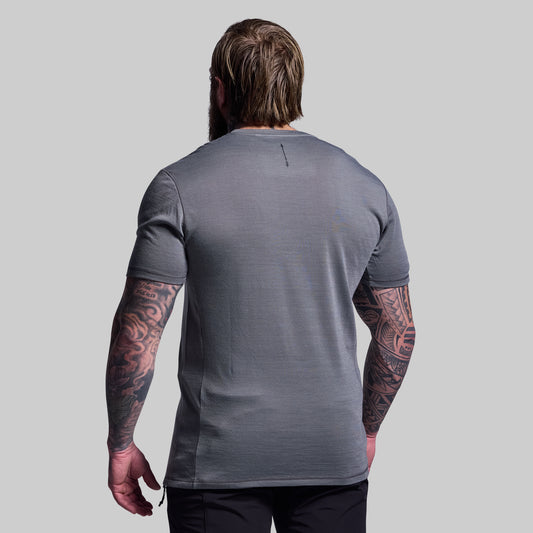 Man wearing a gray t-shirt with visible tattoos on arms against a plain background