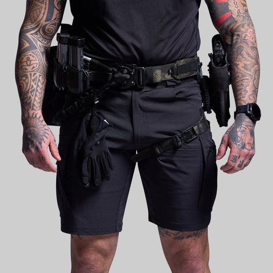Person wearing black tactical shorts and gear with tattoos on arms and legs against a gray background
