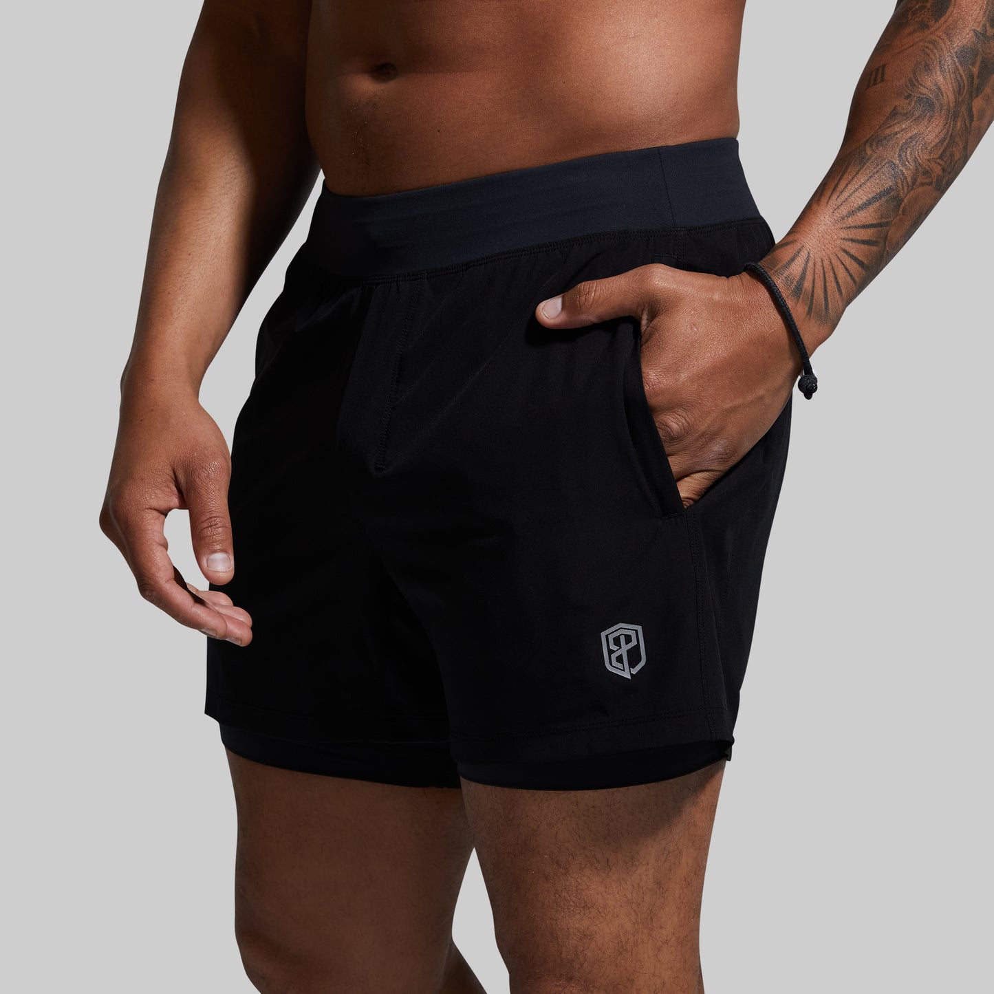 A male model wearing black athletic shorts with pockets and built-in compression shorts, standing with hands in pockets.