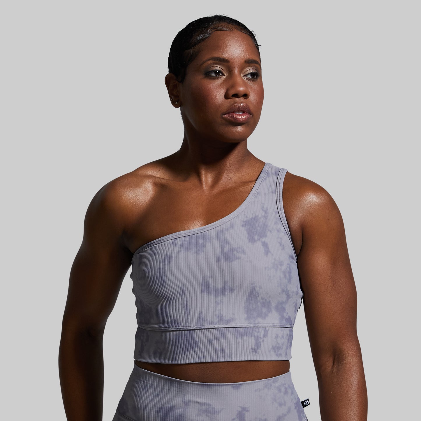 Woman wearing a gray one-shoulder sports bra and leggings against a plain background