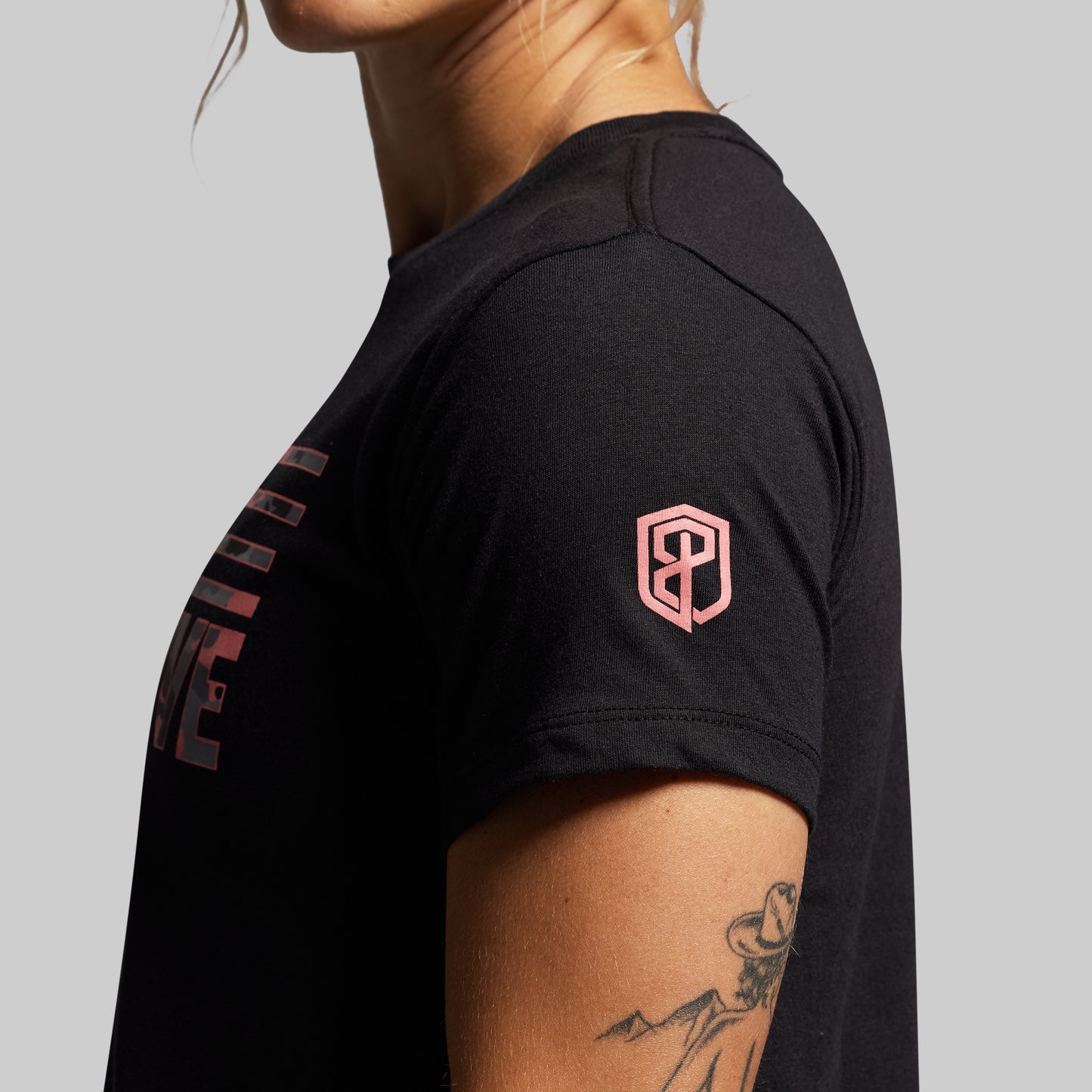 Black t-shirt with a logo on the sleeve and a brand name on the chest, worn by a person against a gray background.