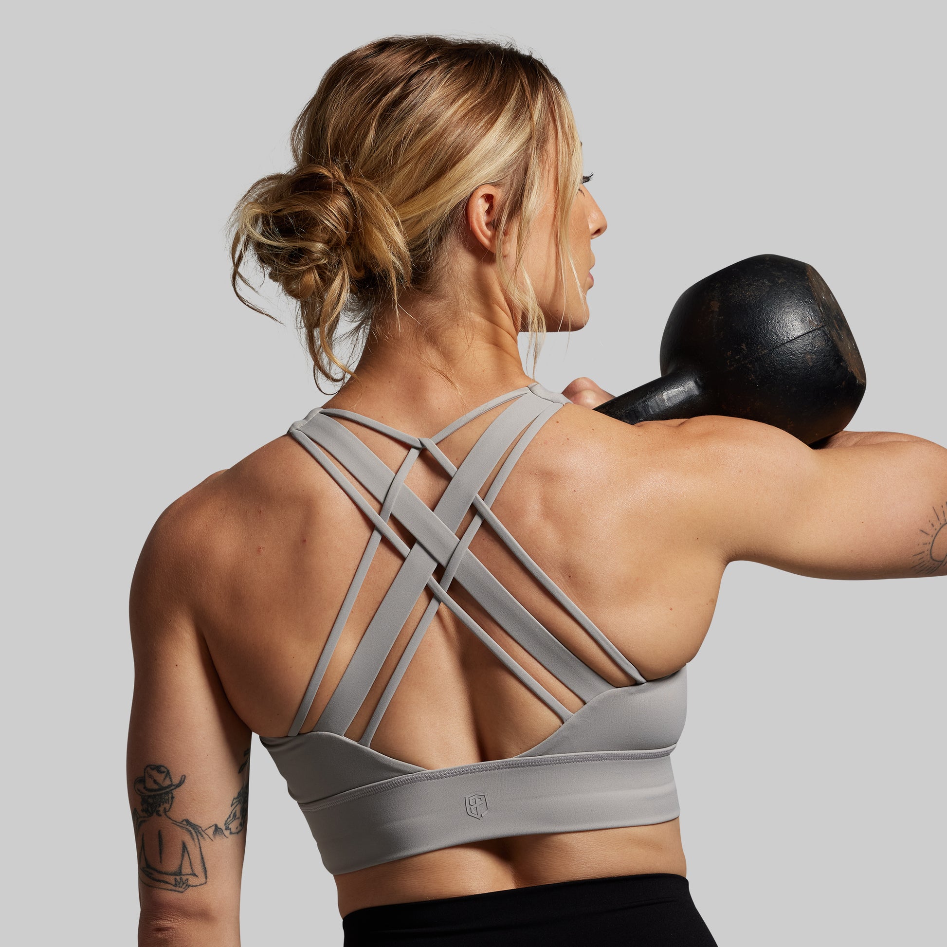 Woman wearing a grey sports bra and lifting a black kettlebell on a gray background