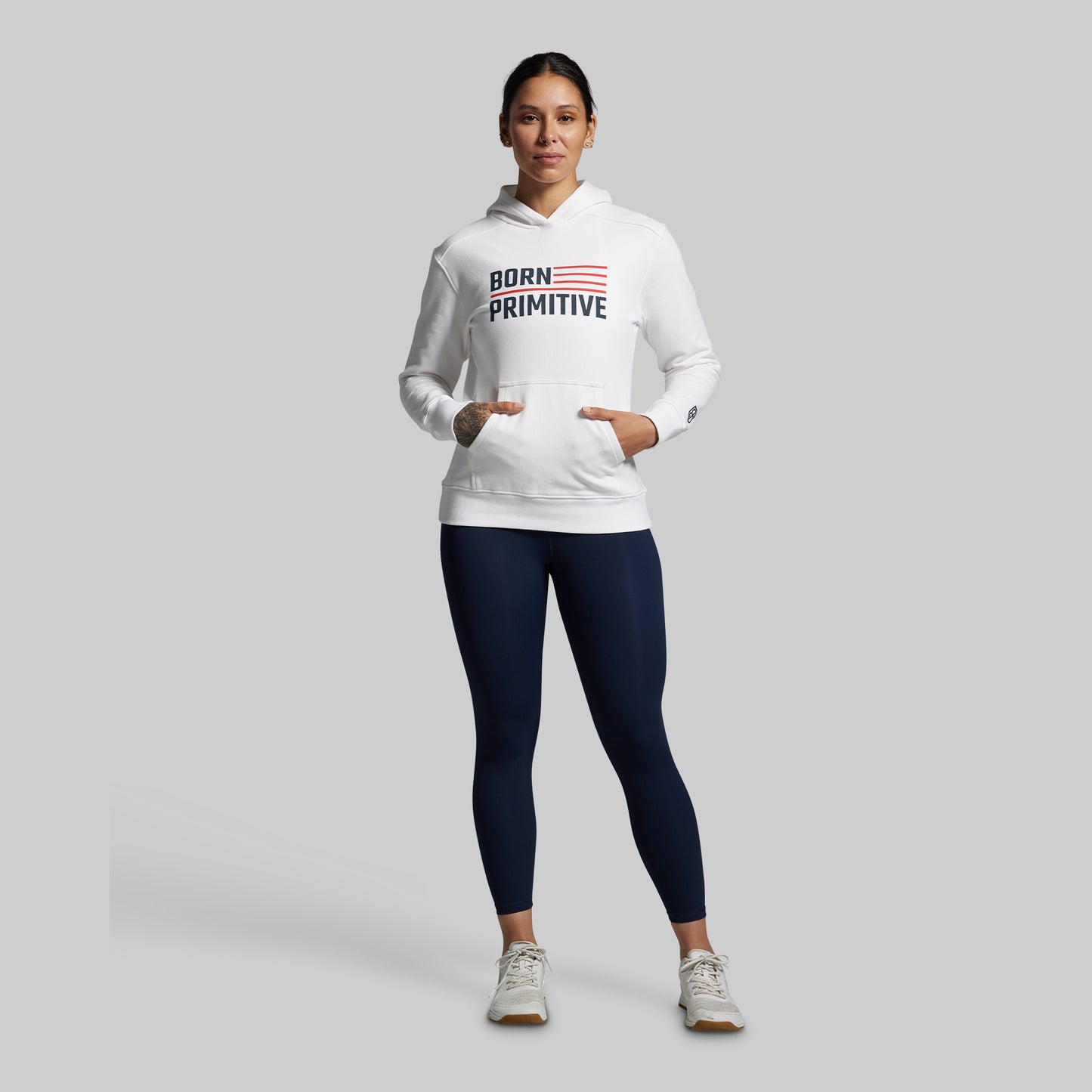 Person wearing a white hoodie with 'Born Primitive' logo and navy leggings on a gray background