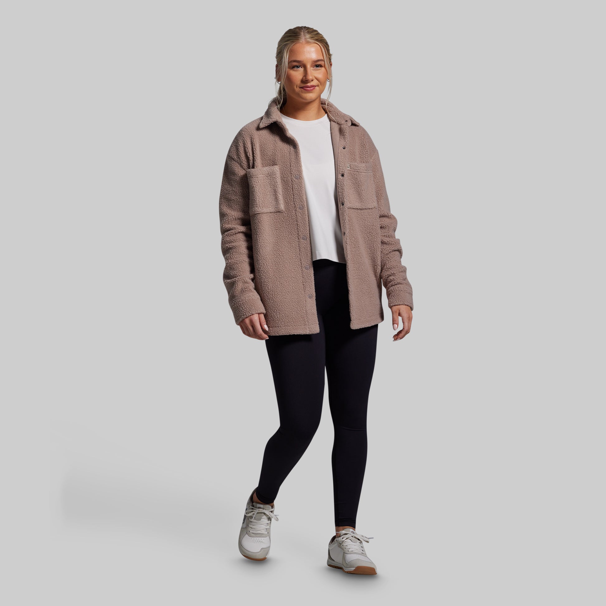 Woman wearing a brown jacket, white shirt, black leggings, and sneakers on a gray background
